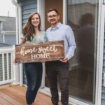 Smiling couple holding 'Home Sweet Home' sign in front of their new house.
